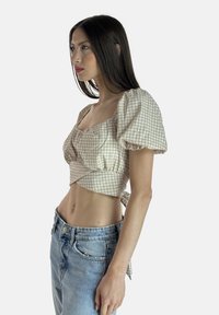 Crop top a scacchi beige e bianchi con maniche a puff corte, scollatura quadrata e design aderente con un fiocco sul retro. Abbinato a jeans azzurri chiari.