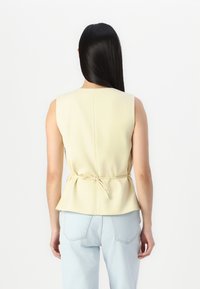 Haut sans manches jaune avec un corsage ajusté et un détail de lien froncé à la taille, associé à un jean en denim bleu clair. Texture du tissu lisse.