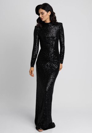 Kobieta o ciemnych włosach ubrana w długą czarną suknię cekinową z długimi rękawami, stojąca na tle jasnoszarym.