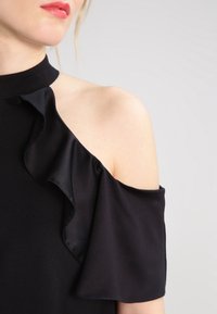 Abito nero senza maniche con colletto alto e dettagli arruffati lungo la spalla, realizzato in un tessuto morbido e liscio.