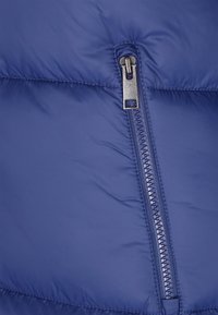 Primo piano di una sezione di giacca imbottita blu con particolare attenzione a una cerniera argentata parzialmente chiusa con dettagli di cuciture visibili.