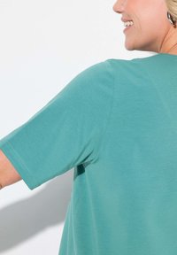 Persona sorridente che indossa una maglietta a maniche corte color turchese, orecchini a cerchio silver, con spalla e profilo laterale visibili su uno sfondo neutro.