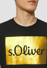 Schwarzes Baumwoll-T-Shirt mit einem großen goldenen rechteckigen Druck, auf dem der Text "s.Oliver" in fetter schwarzer Schreibschrift abgebildet ist. Rundhalsausschnitt.