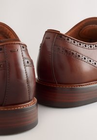 Chaussures en cuir marron avec détails brogues, finition texturée et talon en bois superposé. Bout arrondi et contour lisse à l'arrière.