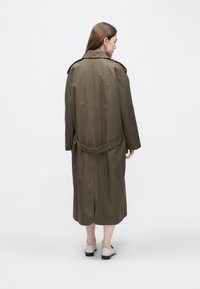 Lång olivgrön trenchcoat med midjebälte, slits bak och axelklaffar, tillverkad av texturerat tyg. Modellen bär ljusa skor.