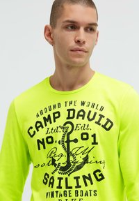 Neongelbes Langarmshirt mit schwarzem Text und Anker-Design, Rundhalsausschnitt und weicher Stoffstruktur.