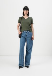 Donna con capelli neri lisci, che indossa una maglietta a V verde oliva, jeans a gamba larga blu, scarpe nere e una piccola borsa a spalla.