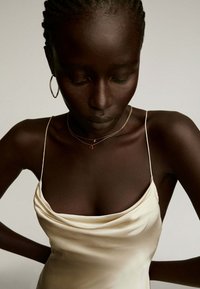 Vestido de satén marfil claro con tirantes finos y escote drapeado, combinado con collares dorados en capas y pendientes de aro. Textura suave, diseño elegante.