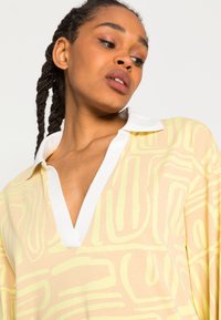 Camisa de manga longa, com decote em V, na cor amarelo claro, com colarinho branco, apresentando padrões abstratos verdes ondulados em tecido leve e suave.
