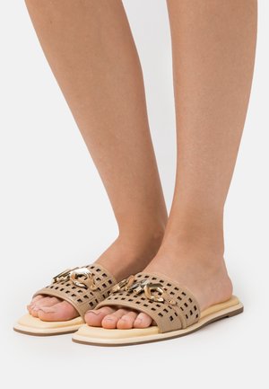 Jalkoja, joissa on beige avonaiset liukumäkikengät, joissa on rei'itetyt hihnat ja kultaketju, seisoo valkoisella pinnalla.