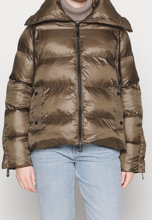 Person trägt glänzende braune Steppjacke mit Frontreißverschluss, strukturierten, geknöpften Taschen, über einem beigen Rollkragenpullover und hellblauen Jeans.
