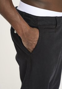 Sort bukser lavet af tekstureret stof med sidelommer; hvid talje strækning delvist synlig øverst. Hånd placeret i den højre lomme.