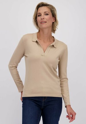 Vrouw met kort blond haar, gekleed in een beige langemouwen polo en donkerblauwe jeans, staand tegen een effen lichte achtergrond.