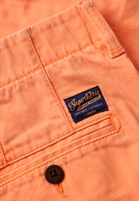 Tessuto arancione con una finitura texturizzata che presenta un'etichetta rettangolare blu con la scritta "Superdry" e un bottone scuro con un dettaglio floreale.