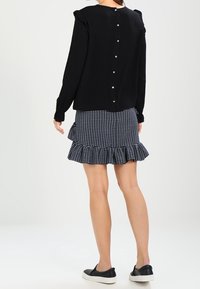 Schwarze Bluse mit langen Ärmeln und gerafften Schultern; Design mit Knopfverschluss hinten. Kombiniert mit einem Rock in Marineblau und Weiß mit einem ausgestellten Rüschensaum.
