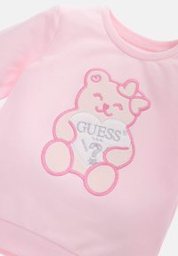 Felpa rosa realizzata in morbido tessuto con un design ricamato di un orso che tiene un cuore. Il cuore presenta la scritta "GUESS" in argento.