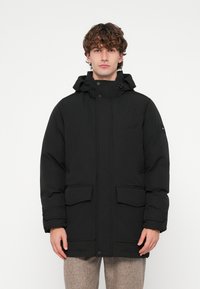 Parka isolada preta com capuz. Possui bolsos frontais grandes e fecho frontal com botões de pressão. Textura lisa com ligeiro brilho.