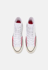 Høytopp sneakers laget av hvit kanvas med røde detaljer, svart kant, og en gummitåhette. Har hvite lisser og metalløyer.
