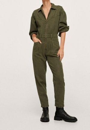 Vrouw draagt een olijfgroene jumpsuit met rits, opgerolde mouwen en zwarte enkellaarzen, staat met één hand in de zak.
