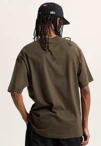 Camiseta de algodón verde oliva con mangas cortas, corte relajado y escote redondo; combinada con una gorra negra con acentos de logo rosa.