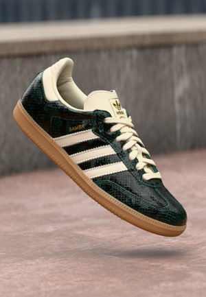 Ciemnozielony, teksturowany but sportowy Adidas Samba z kremowymi sznurówkami, białymi paskami i gumową podeszwą, unoszący się nad różowawym podłożem.