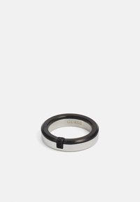 Silver and black ring with a smooth finish, featuring a thin black band and a small accent detail on the side. Engraved "GUESS" inside.