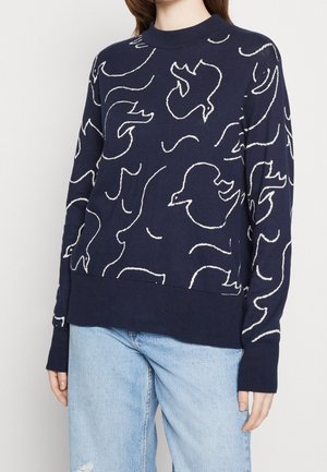 Person mit einem marineblauen Pullover mit weißen abstrakten Vogelmustern und hellblauen, zerrissenen Jeans, stehend vor einem schlichten, hellen Hintergrund.