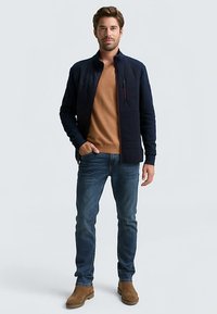 Marineblaue gesteppte Jacke mit Fronttaschen und rotem Reißverschlussdetail, getragen über einem hellbraunen Pullover und blauen Jeans, ergänzt durch braune Schuhe.