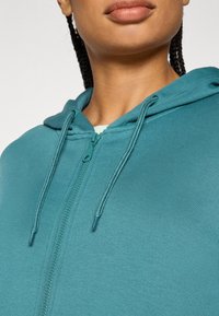 Τζελ κουκούλα με φερμουάρ σε τιρκουάζ χρώμα, με απαλό  ύφασμα, ρυθμιζόμενα κορδόνια και μπροστινό φερμουάρ. Οι λεπτομέρειες κοντινής απόστασης τονίζουν το ράψιμο και την ποιότητα του υφάσματος.