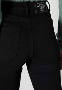 Černé vysoké džíny s fitted siluetou, s dvěma zadními kapsami a štítkem na pásku, který označuje 30. výročí.