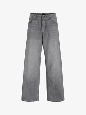 Ung person står, iført svart hettejakke med glidelås, hvit t-skjorte, løse grå jeans og svarte sko, ser litt til siden med et smil.