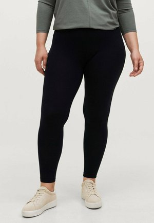 Schwarze Leggings aus dehnbarem Stoff mit einer glatten Textur und einer figurschmeichelnden Passform, kombiniert mit einem grauen Oberteil und beigen Turnschuhen.