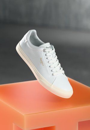 White low-top sneaker with laces and gold “Pepe Jeans London” logo, displayed above an orange cube on gray background.