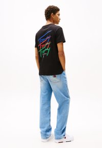 Black cotton t-shirt featuring colorful graphic text on the back. Paired with light blue denim jeans and white sneakers.