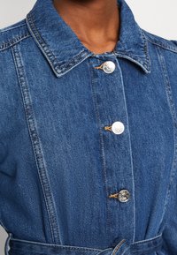 Giacca di jeans con colletto, chiusura con bottoni argentati e vita con cintura. Presenta un lavaggio blu medio e una texture di cucitura visibile.