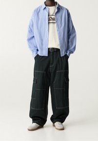 Chemise à rayures bleues à boutons, t-shirt graphique crème, pantalons cargo foncés avec poches, et baskets grises avec semelles marron. Accessoire chaîne argentée.