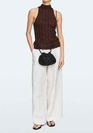 Femme portant un top marron sans manches en maille côtelée, un pantalon large blanc, un sac bandoulière noir tressé et des sandales tongs noires, debout les mains détendues.