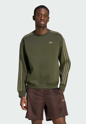 Jonge man draagt een olijfgroene Adidas trui met strepen op de mouwen en bruine Adidas shorts met logo op het linkerbeen.