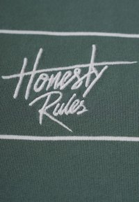 Grüner Stoff mit einer gerippten Textur, versehen mit weißem gesticktem Text, der "Ehrliche Regeln" liest, und einem horizontalen weißen Streifendetail.