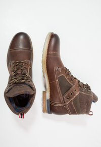 Botas de cuero marrón hasta el tobillo con acentos de fieltro beige, costuras texturizadas y una suela de goma transparente. Presenta cordones y ojales de metal.