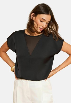 Woman wearing a black short-sleeve top with a sheer V-neck panel, white high-waist pants, and a gold bracelet on her left wrist.