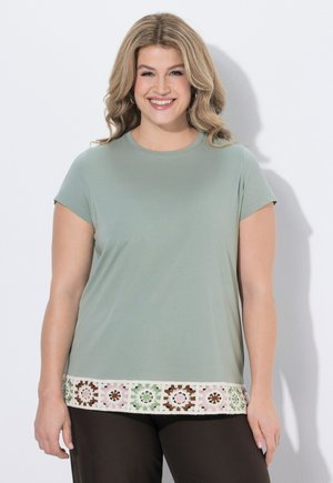 Lächelnde Frau mit welligem blondem Haar, die ein salbeigrünes Kurzarmshirt mit mehrfarbigem Häkelbesatz und dunkle Hosen trägt, steht vor einer weißen Wand.