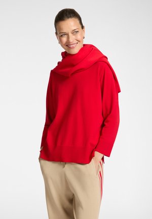 Donna sorridente, che indossa un maglione rosso brillante con una sciarpa coordinata drappeggiata sulle spalle e pantaloni beige con strisce rosse, mani nelle tasche.