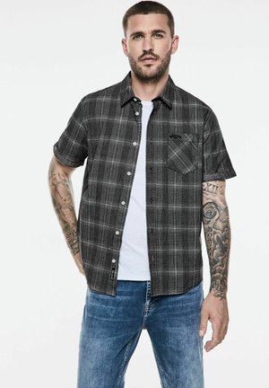 Kurzärmeliges Karoshemd in Schwarz und Weiß, mit Knopfleiste und Tasche. Über einem weißen T-Shirt getragen, kombiniert mit blauen Jeans.