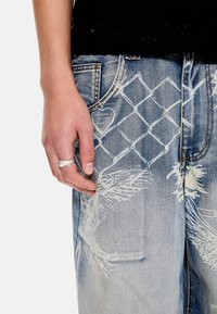 Αριστερό χέρι με ασημένιο δαχτυλίδι ακουμπισμένο δίπλα σε τζιν τζιν με διακοσμήσεις λευκών σχεδίων αλυσίδας και φτερών.