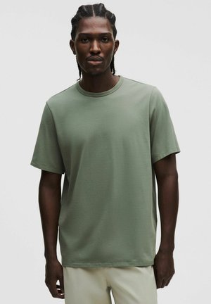 Mężczyzna stojący z warkoczami, ubrany w prostą oliwkowozieloną koszulkę z krótkim rękawem i jasne spodnie na neutralnym tle.