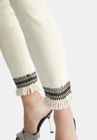 Pantalones de mezclilla blancos con un borde de encaje negro con flecos en el dobladillo. Tejido suave con un diseño de pierna recta. Llevados con sandalias negras de tacón.