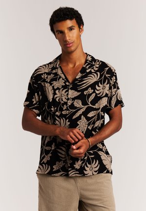 Mann passt Perlenarmband an, trägt schwarzes kurzärmeliges Hemd mit beiger tropischer Blattmusterung und beige strukturierte Hose, neutraler Hintergrund.