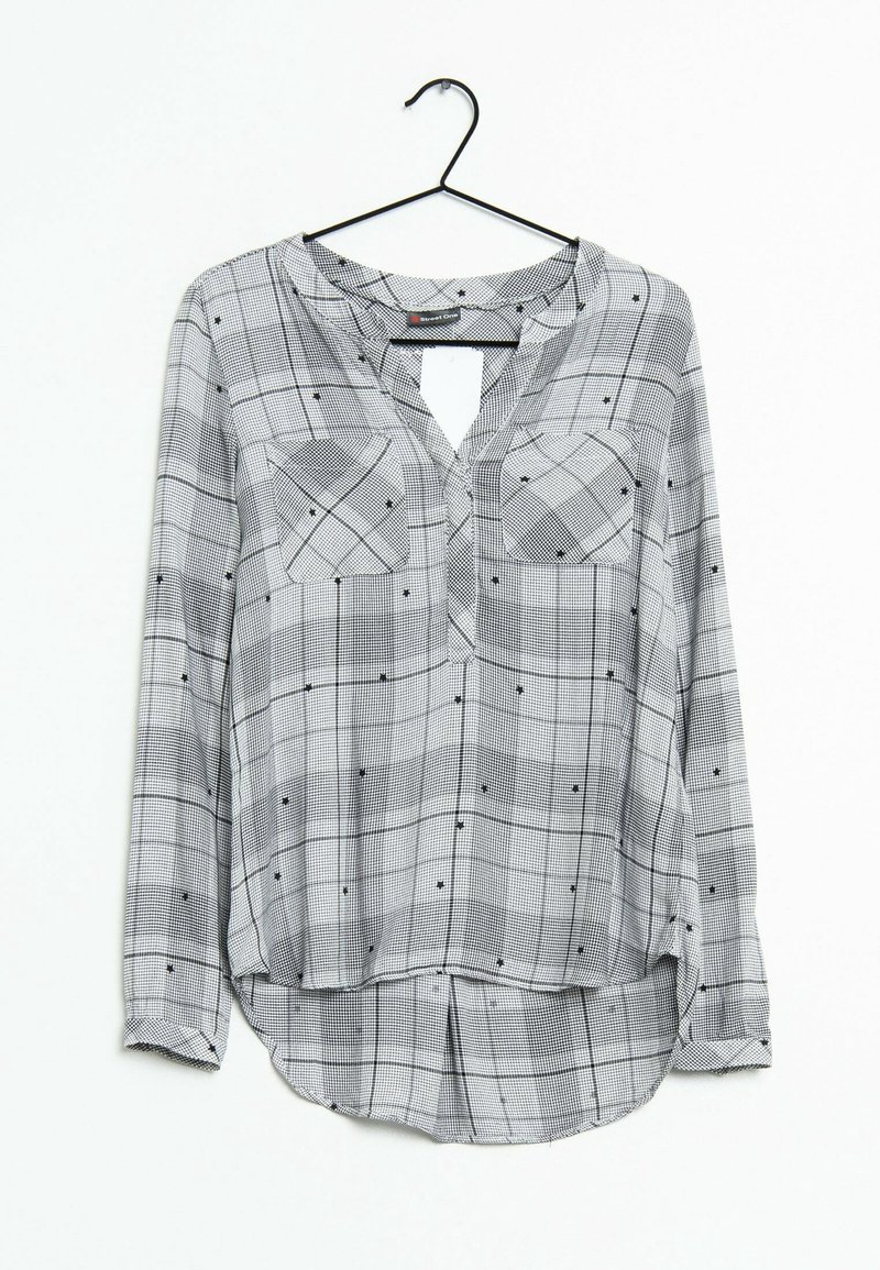 Blouse femme à manches longues avec motif à carreaux noir et blanc, poches avant et col en V, suspendue sur un cintre noir sur fond blanc.