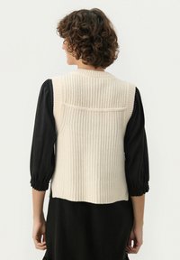 Brede gebreide vest in cr�ème met een textuur ribpatroon, voorzien van een hoge kraag en open achterkant, gecombineerd met een zwarte blouse met pofmouwen eronder.